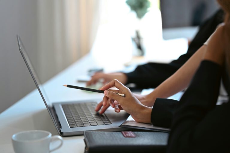 Two people sit side by side at a white desk, collaborating on Carlisle web design. One points at the laptop screen with a pencil while the other types. A white coffee cup and closed black notebook rest on the softly blurred background.