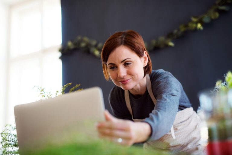 A woman with short brown hair, wearing a grey top and a white apron, smiles and leans towards a laptop, perhaps working on Carlisle web design. She is surrounded by green plants and soft window light, with a dark wall and leafy garland behind her.
